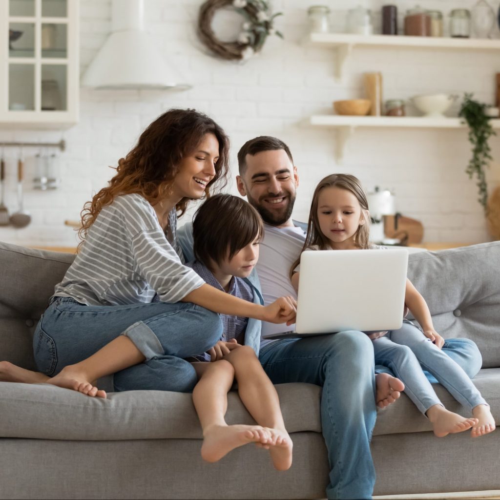 Happy young family with little kids sit on sofa in kitchen have fun using modern laptop together, smiling parents rest on couch enjoy weekend with small children laugh watch video on computer at home